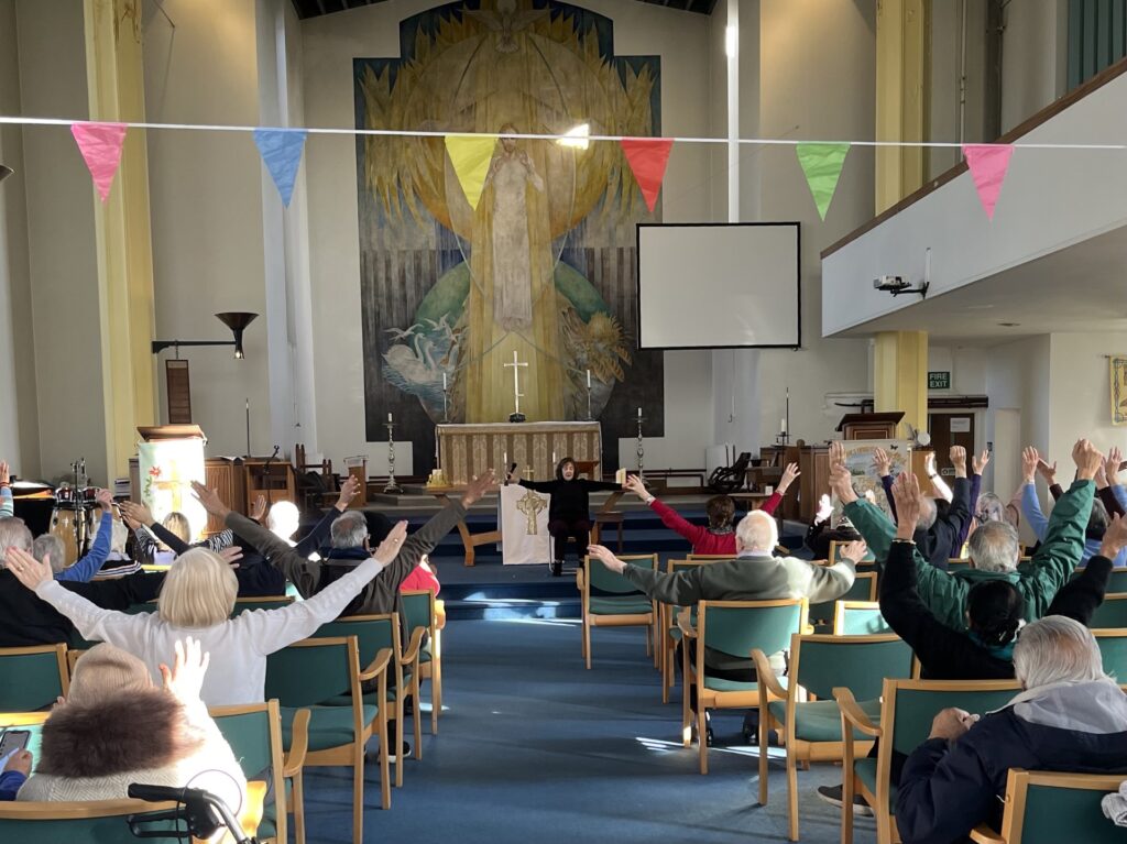 A lady at the front of a church leads chair-based exercises. A group of elderly people are seated facing her and are following the exercises. Their arms are outstretched in the air.