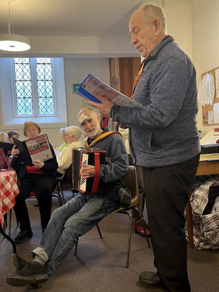 A man leading a sing-along is standing looking at a songbook. Another man is seated and playing the accordion. In the background, others looks at their own songbooks.