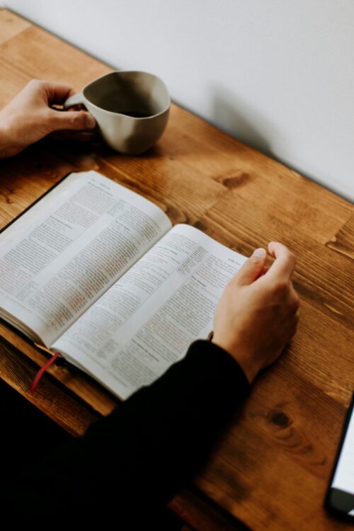 A close up of two hands on a desk - one holds a coffee cup and the other rests on a Bible.