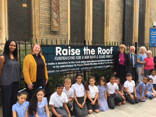 Gathering of small children in front of a sign labeled 'raise the roof - fundraising for a new roof and solar panels'. on either side of the sign stands the vicar and the bishop of london alongside congregation members.