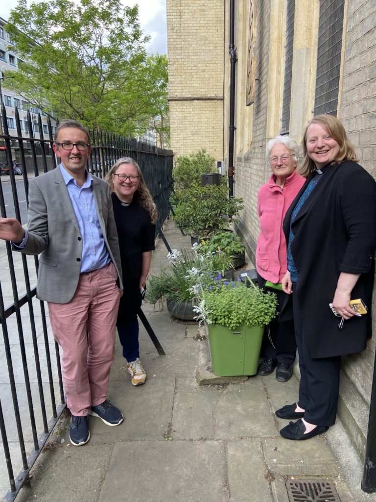 The St Paul's Marylebone Eco team - a smiling man in grey suit, two women in clergy collars, one older female volunteer. all smiling. standing in front of a number of potted plants outside the church.