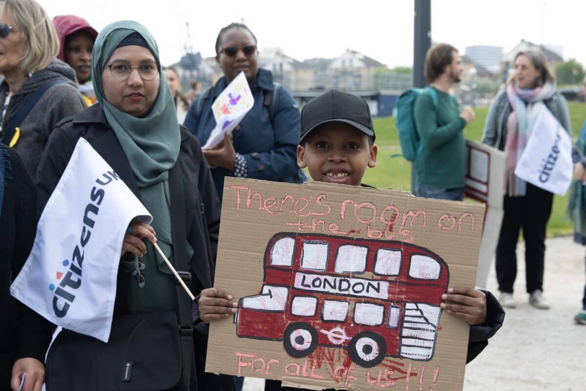Boy holding up a cardboard sign saying "There's room on the bus for all of us", with woman in headscarf standing next to him wtih a Citizens flag. Others in background as part of an organised gathering.