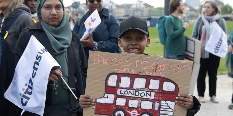 Boy holding up a cardboard sign saying "There's room on the bus for all of us", with woman in headscarf standing next to him wtih a Citizens flag. Others in background as part of an organised gathering.