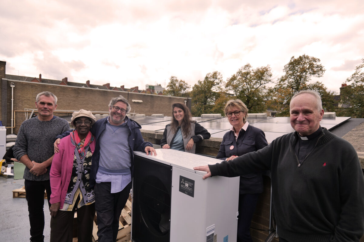 A group of smiling people standing next to an air source heat pump, including a vicar with dog collar
