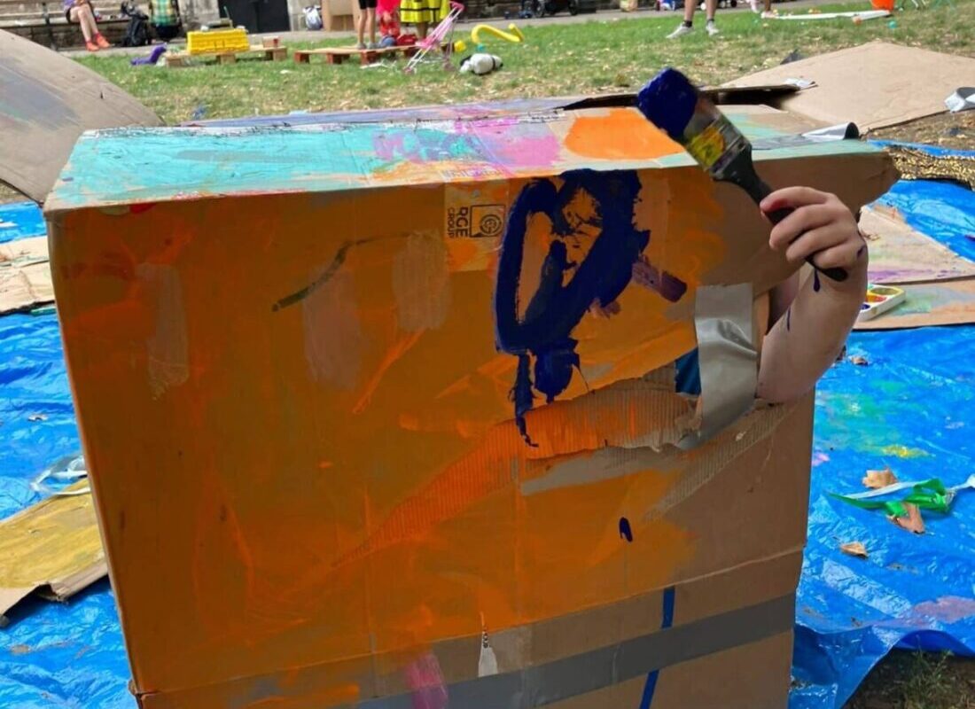 The image shows a large, painted cardboard box. A child's arm is poking out of it holding a paintbrush.