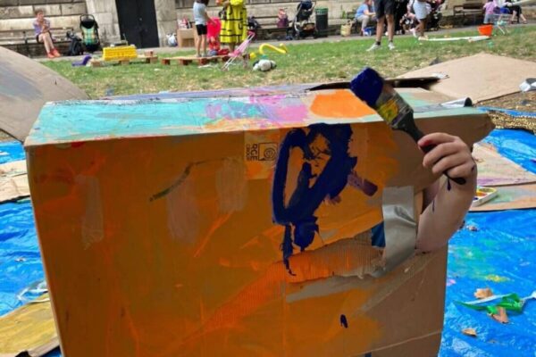 The image shows a large, painted cardboard box. A child's arm is poking out of it holding a paintbrush.