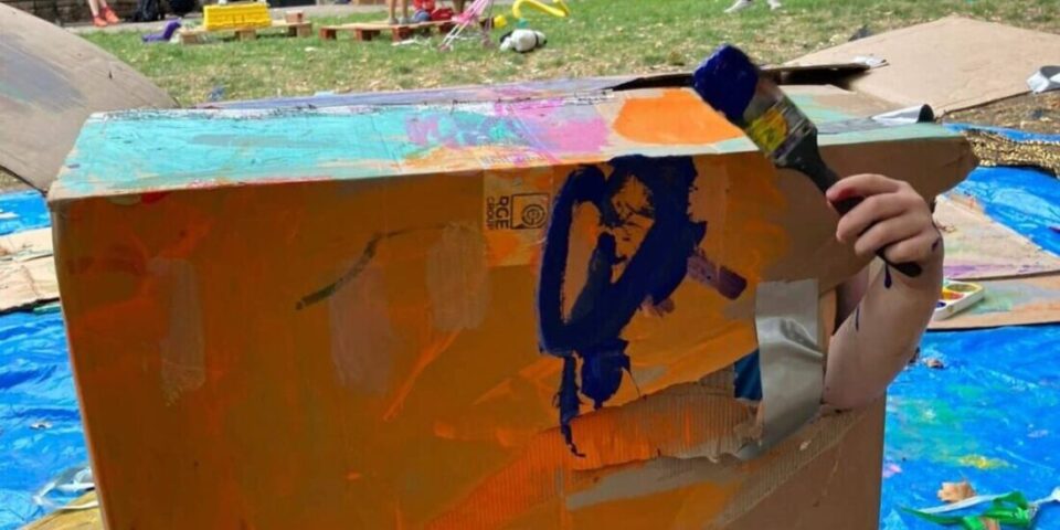 The image shows a large, painted cardboard box. A child's arm is poking out of it holding a paintbrush.