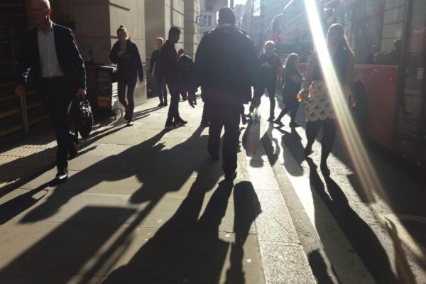 a bustling london street with bright sun and shadows of pedestrians, alongside a London bus